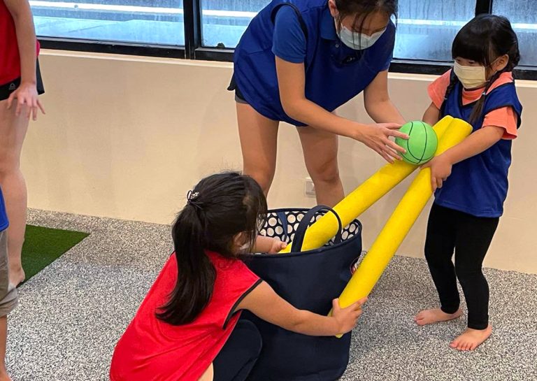 兩位幼兒合作使用浮力棒夾住球移動,這項趣味競賽旨在訓練團隊合作 (Teamwork) 精神與物體操控 (Object Control) 能力,提升孩子溝通與協調的社交技巧。