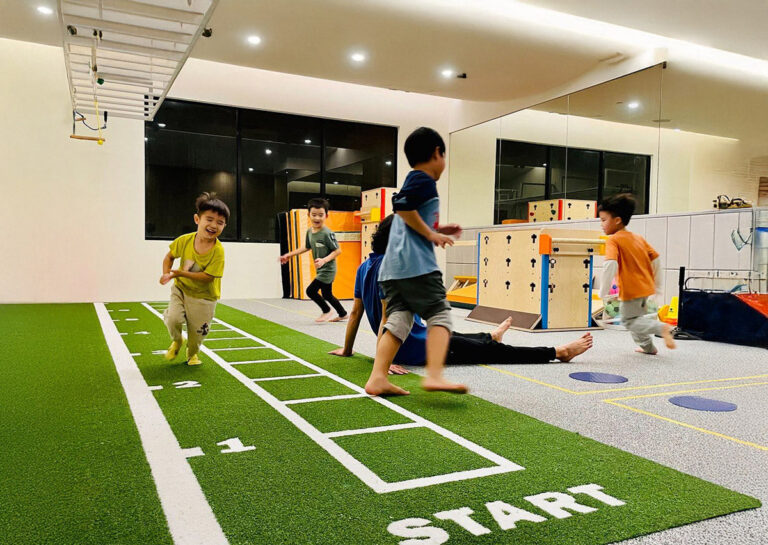 兒童在室內草皮跑道上快樂奔跑，訓練位移技巧 (Locomotor Skills) 與腿部爆發力，透過團體追逐遊戲提升心肺耐力與運動樂趣。