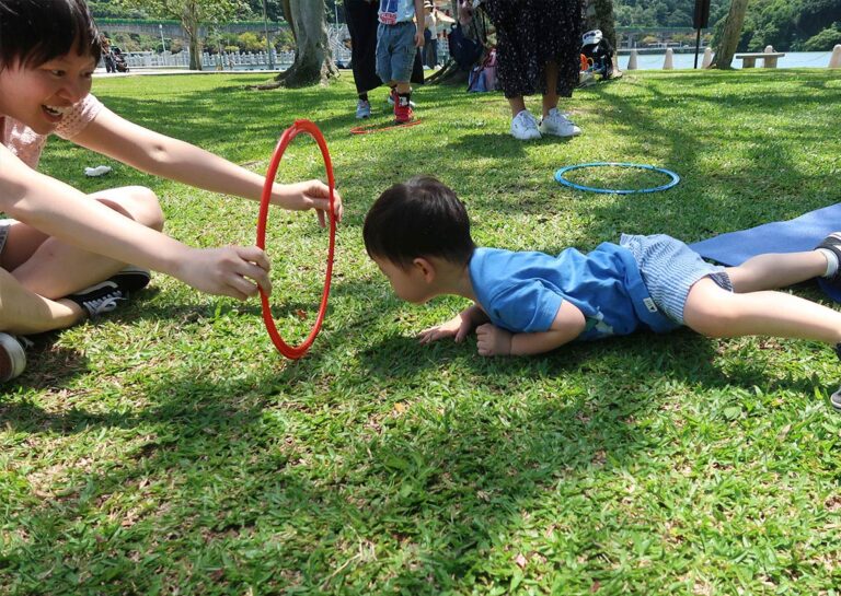 幼兒在戶外草地上進行匍匐爬行穿越呼拉圈,草地的質感能提供豐富的觸覺刺激 (Tactile Stimulation),爬行動作則能有效強化核心肌群與肩帶穩定度。