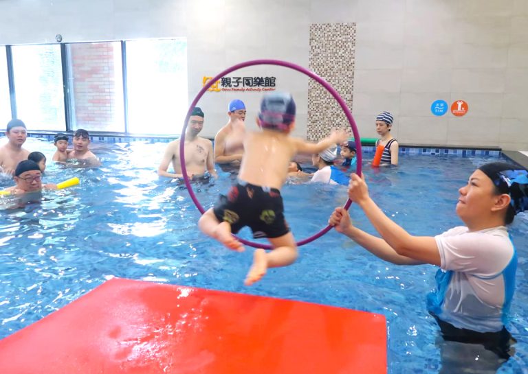 穿著 Oovi 制服的教練手持呼拉圈,引導兒童從浮墊上勇敢跳水 (Jumping Entry) 穿越圓圈,透過遊戲化教學 (Gamification) 訓練入水膽量與空間判斷力。