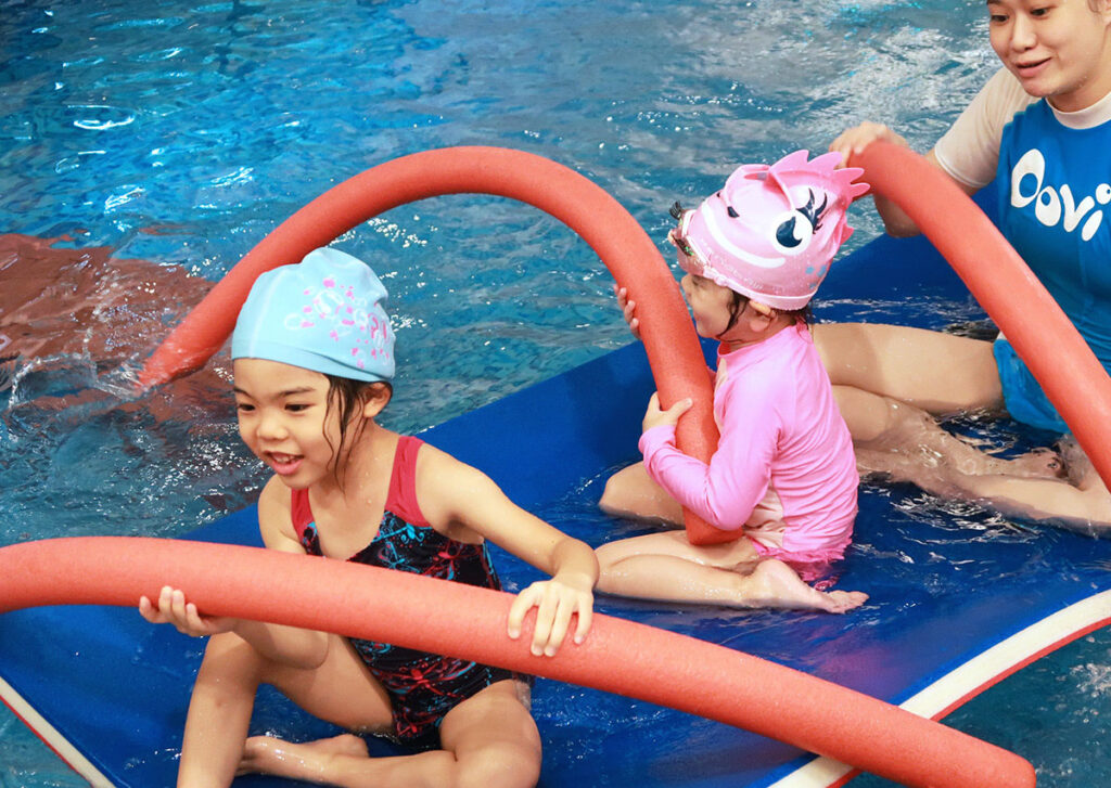 幼兒坐在藍色浮墊上將浮力棒當作船槳,進行划船遊戲 (Rowing Game),這種遊戲化教學 (Gamification) 能有效訓練幼兒的上肢協調性與核心穩定度。