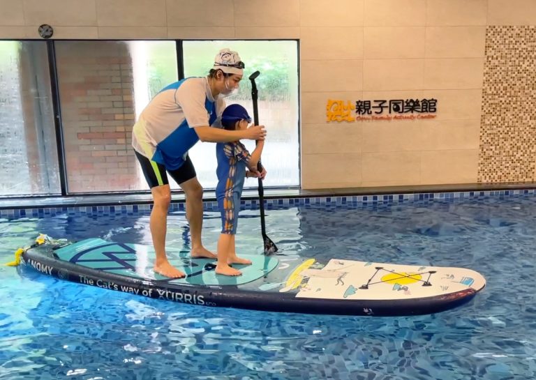 穿著 Oovi 制服的專業教練指導兒童在室內泳池進行水上立槳 (SUP) 訓練,引導孩子在不穩定的板上維持動態平衡 (Dynamic Balance) 並操作划槳。這項特色課程能同時強化核心肌群、專注力與全身協調性。