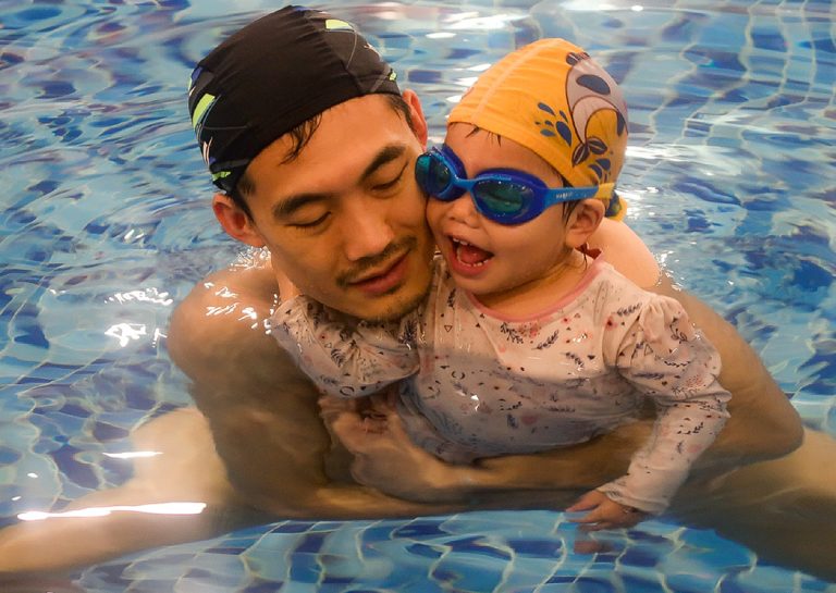爸爸抱著配戴泳鏡的幼兒在水中親密互動,透過正向鼓勵讓孩子習慣穿戴游泳裝備,並享受親子共游的快樂時光。