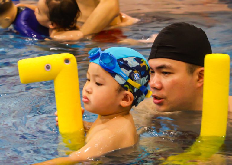 爸爸陪伴幼兒使用黃色浮條進行打水練習,透過教具輔助建立水中浮力認知,並強化親子共游的信任感與安全感。