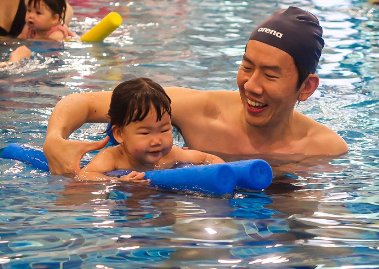 爸爸與孩子在泳池中使用藍色浮條互動大笑,透過遊戲化學習 (Play-based Learning) 讓孩子對游泳產生正向連結,降低學習壓力。
