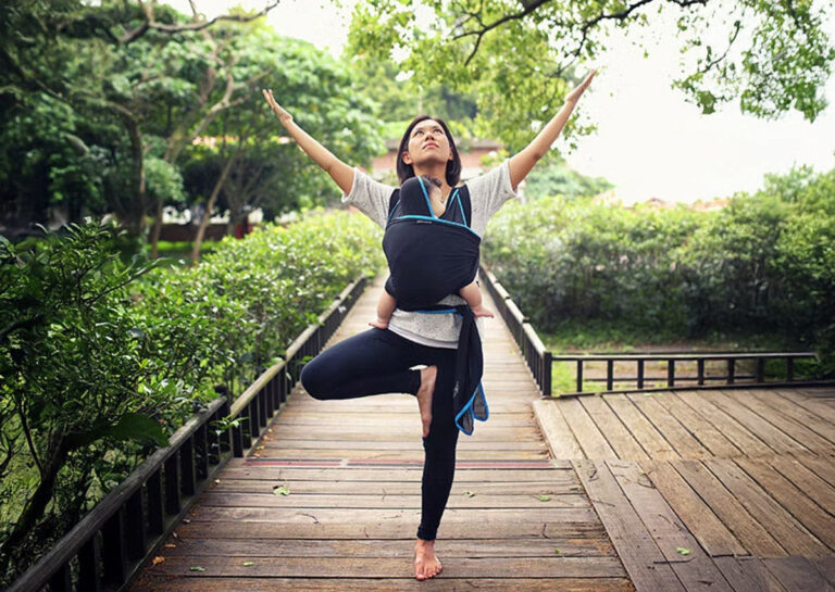 媽媽揹著寶寶在戶外進行揹巾瑜珈 (Babywearing Yoga)，展示揹巾穿戴能結合產後運動，幫助媽媽找回身心平衡與核心力量，享受與寶寶的寧靜時光。