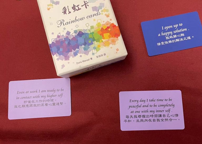 Oovi 揹巾穿戴師資培訓:彩虹卡 (Rainbow Cards) 與內在指引同學分享。「彩虹卡 (Rainbow Cards)」外盒與三張示例卡片陳列於紅色桌面上。卡片上印有中英文對照的心靈指引短句,例如藍色卡片上的「我敞開心胸接受快樂的解決之道」,用於輔助學員進行內在覺察與情緒引導。