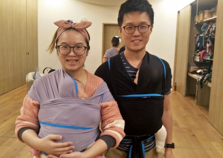 夫妻同時使用彈性揹巾 (Stretchy Wrap) 揹負新生兒,展示共同育兒 (Co-parenting) 的美好畫面。透過這類柔軟材質的揹巾,能提供如袋鼠式護理般的包覆,建立全家人的親密依附關係。