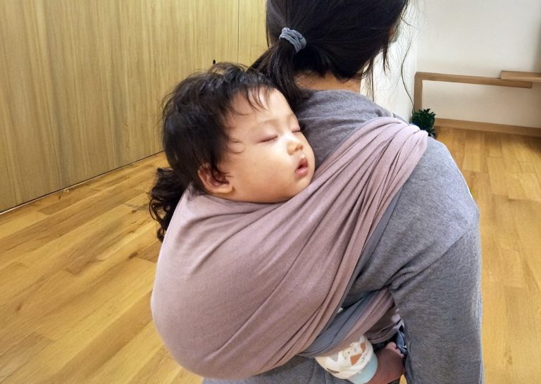 幼兒在媽媽背上使用揹巾進行後揹 (Back Carry) 並安穩熟睡,展示揹巾能提供極佳的包覆感與安全依附 (Secure Attachment),是安撫高需求寶寶入睡與家長解放雙手的完美解決方案。