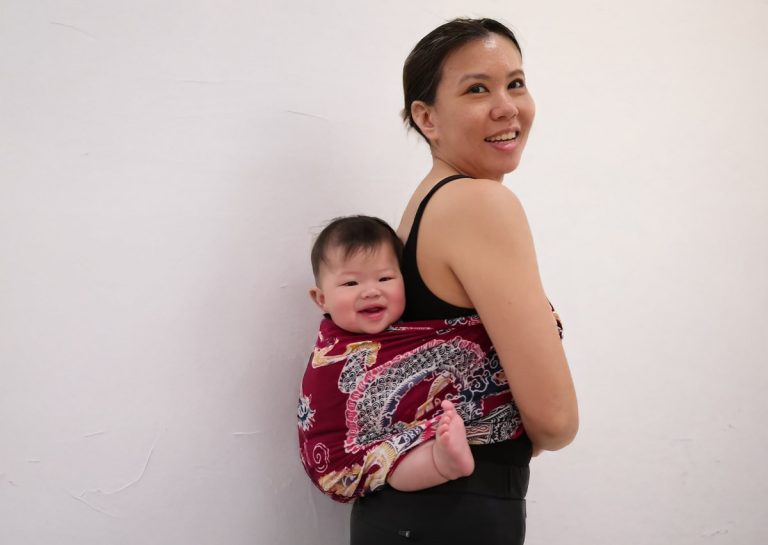 媽媽自信展示紅色圖騰的編織長布揹巾 (Woven Wrap),呈現揹巾穿戴不僅符合人體工學舒適度,更能成為展現家長個人風格的時尚配件,打破育兒用品單調的刻板印象。