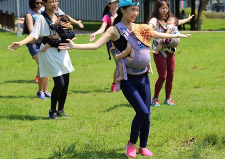 媽媽們在戶外草地上揹著寶寶進行揹巾律動 (Babywearing Dance),透過伸展動作找回產後核心力量與身體平衡,同時讓寶寶在媽媽懷中享受大自然的感官刺激。