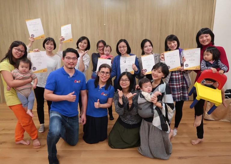 Oovi 揹巾穿戴5.C.M.師資培訓結業合影:見證專業與溫度的起點。多位女性學員手持國際認證證書,懷中抱著嬰幼兒,展現結訓的喜悅與專業成果。前方身著 Oovi 藍色制服的講師比出讚的手勢,背景為木質溫馨教室。