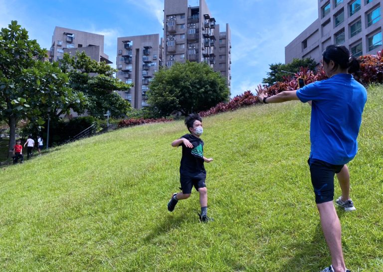 孩童在 Oovi 暑假營隊的戶外綠地向教練奔跑挑戰坡度,透過自然地形的移動訓練鍛鍊下肢力量、心肺耐力與環境適應力。